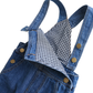 Close-up of a blue denim overall with adjustable straps, gold-tone snap buttons, and a black-and-white gingham lining visible at the neckline, photographed on a white background.