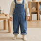A toddler standing indoors wearing blue denim overall-style pants over a cream and navy striped long-sleeve shirt, with cream socks. The room has a light wood floor, a small wooden table, and a wooden shelf in the background.