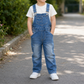 A child standing on a paved path outdoors, wearing blue denim overall with a white t-shirt and white sneakers.